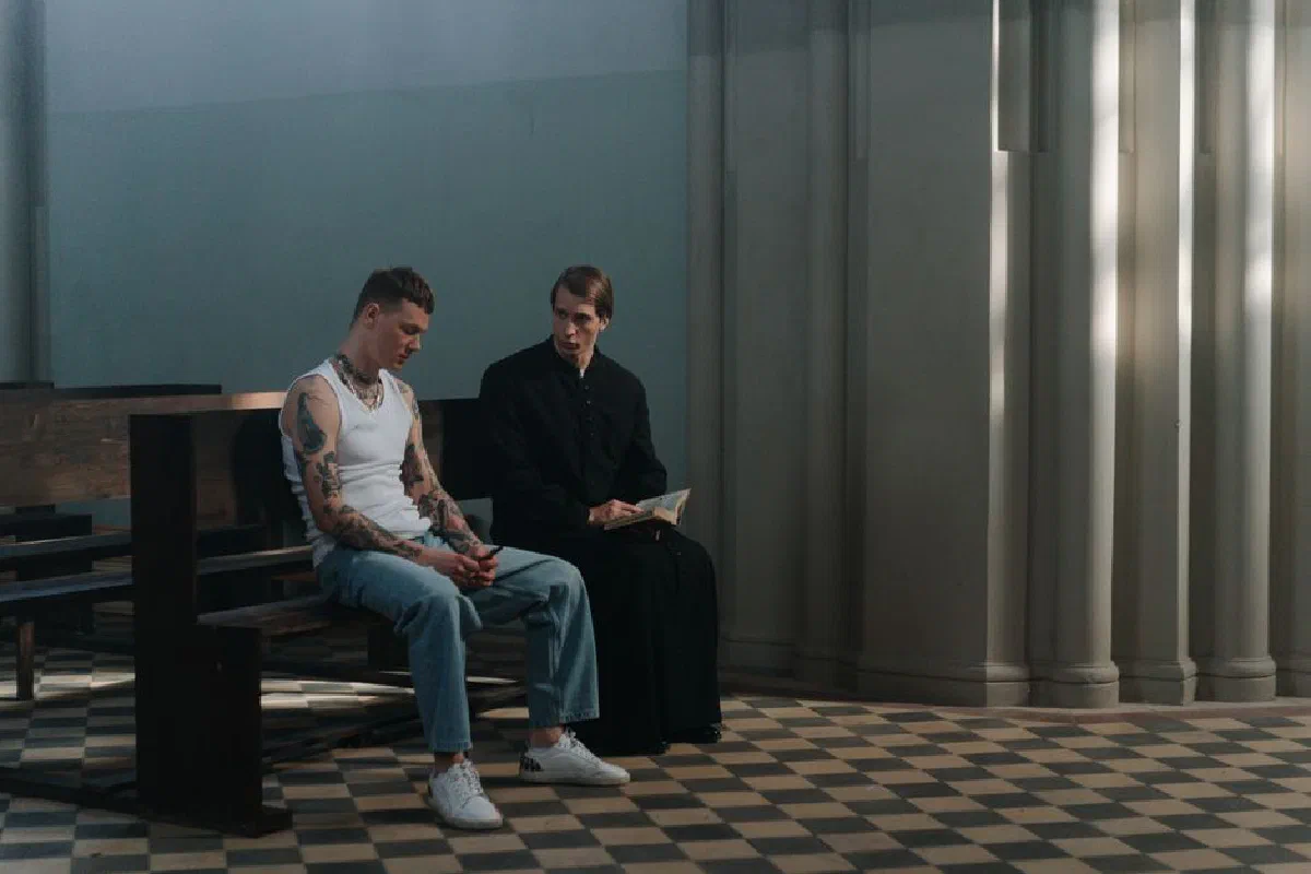 A tattooed man seeks counsel from a priest in a serene church interior, highlighting themes of faith and reflection.