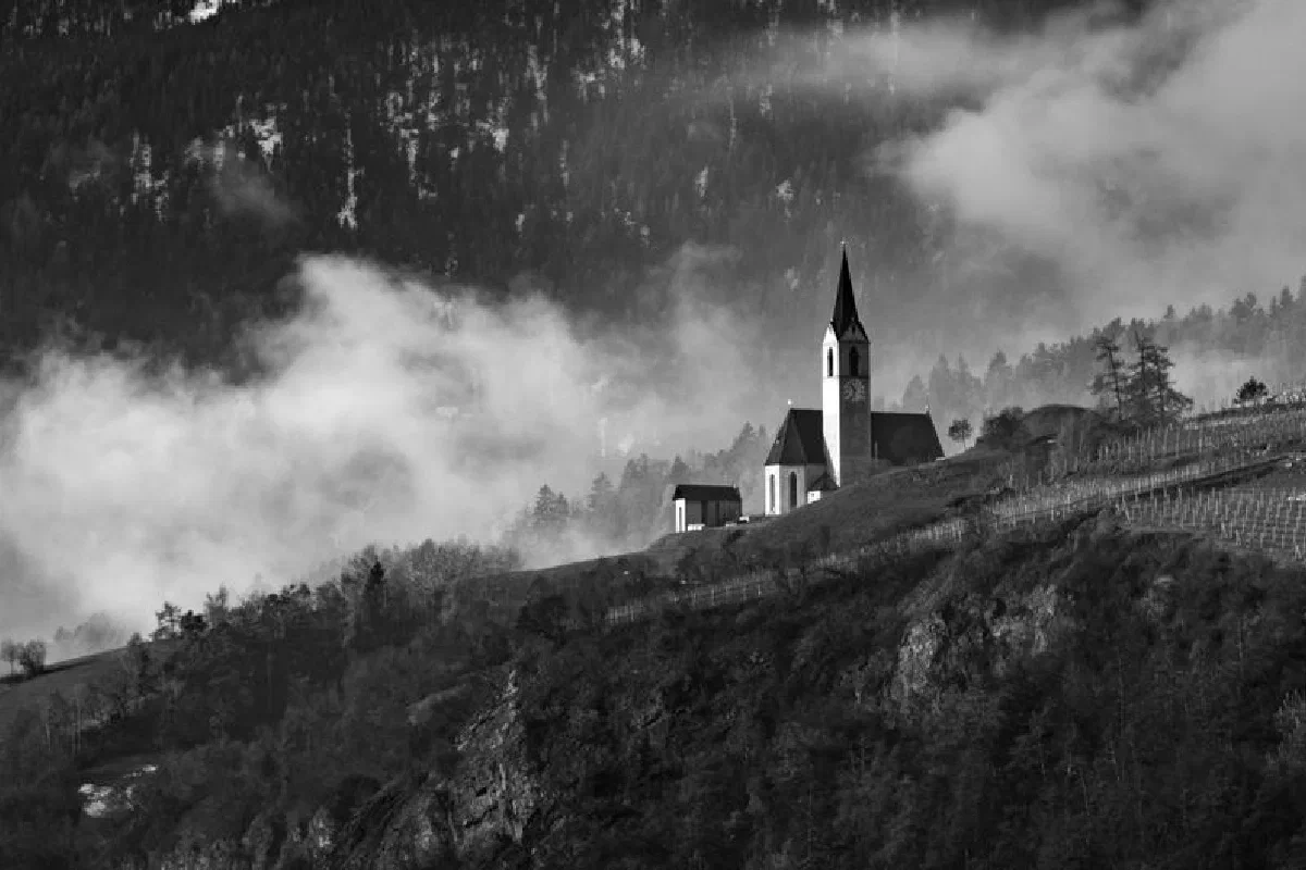 Black and white photo of a church on a misty hillside with mountains in the background.