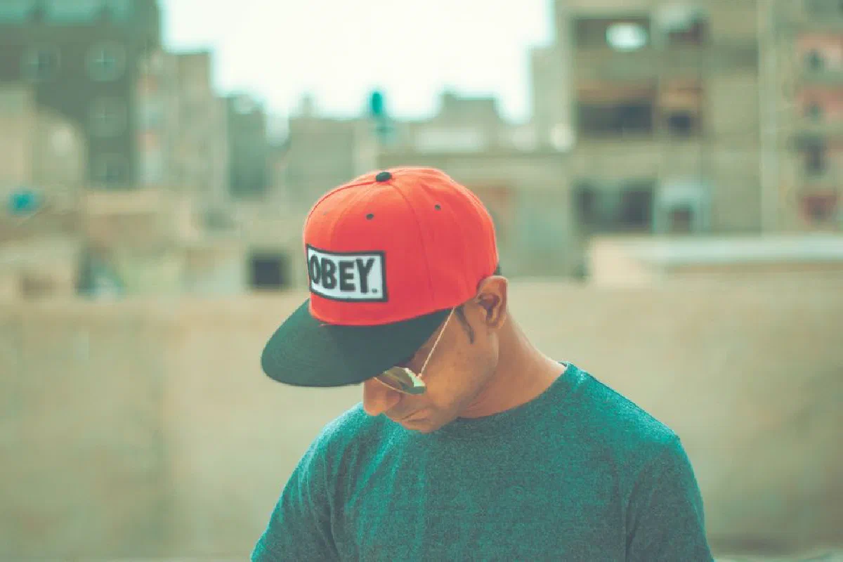 Casual portrait of man wearing sunglasses and red Obey cap, looking down outdoors.