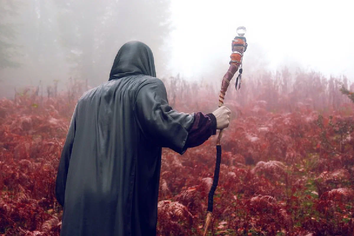A cloaked figure in a fantasy setting walks through a foggy, red-tinged forest holding a magical staff.