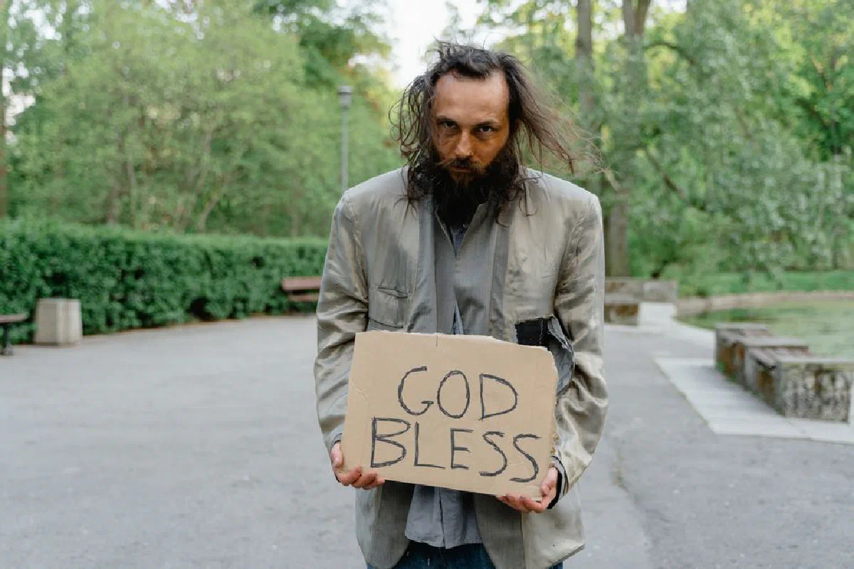 Portrait of a homeless man holding a 'God Bless' sign in a park setting.