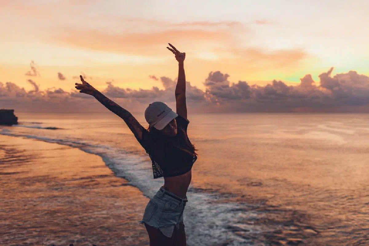 Side view fit lady in casual shorts and cap enjoying evening on seashore while standing with arms raised against calm endless sea and majestic bright cloudy orange sky during sunset