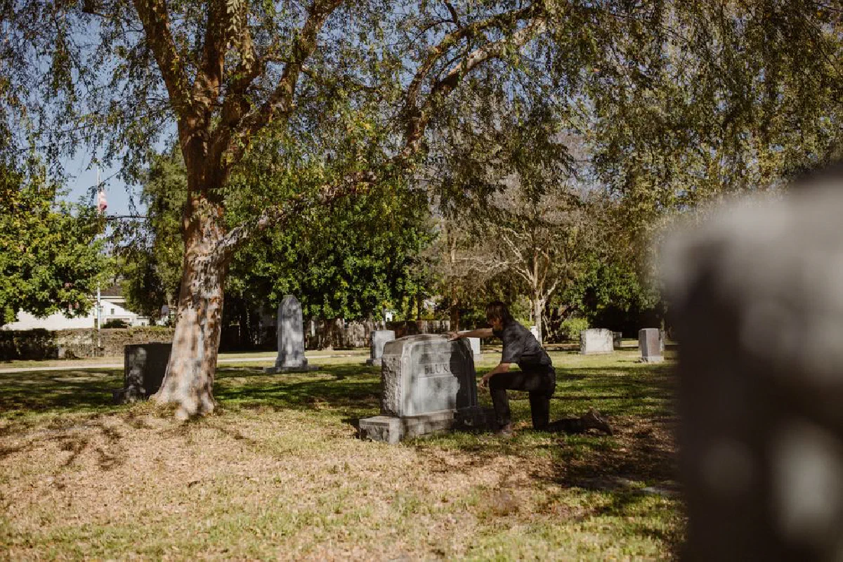 A man kneels by a grave in a sunlit cemetery, reflecting moments of grief and solitude.