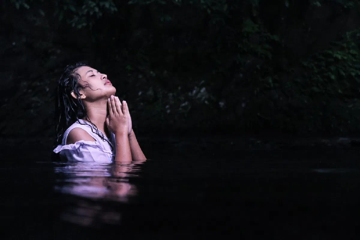 A woman in water amidst lush greenery, hands clasped in prayer under twilight.