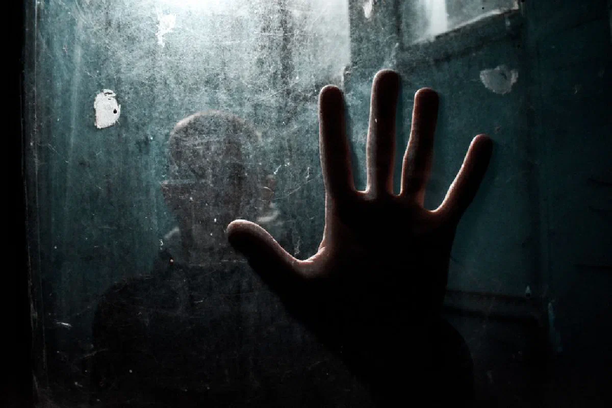Silhouette of a hand pressed against a dirty, moody glass panel with a person faintly visible.
