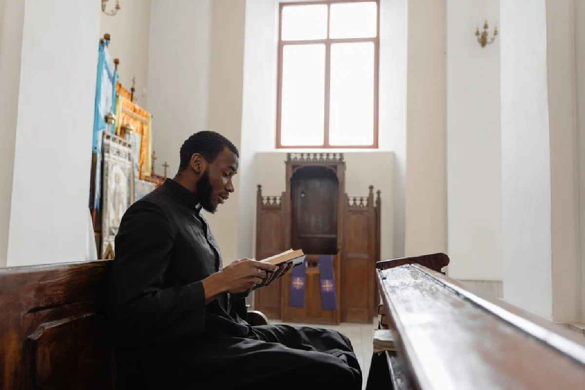 An African American priest sitting in a church pew, reading a book in a serene church interior.