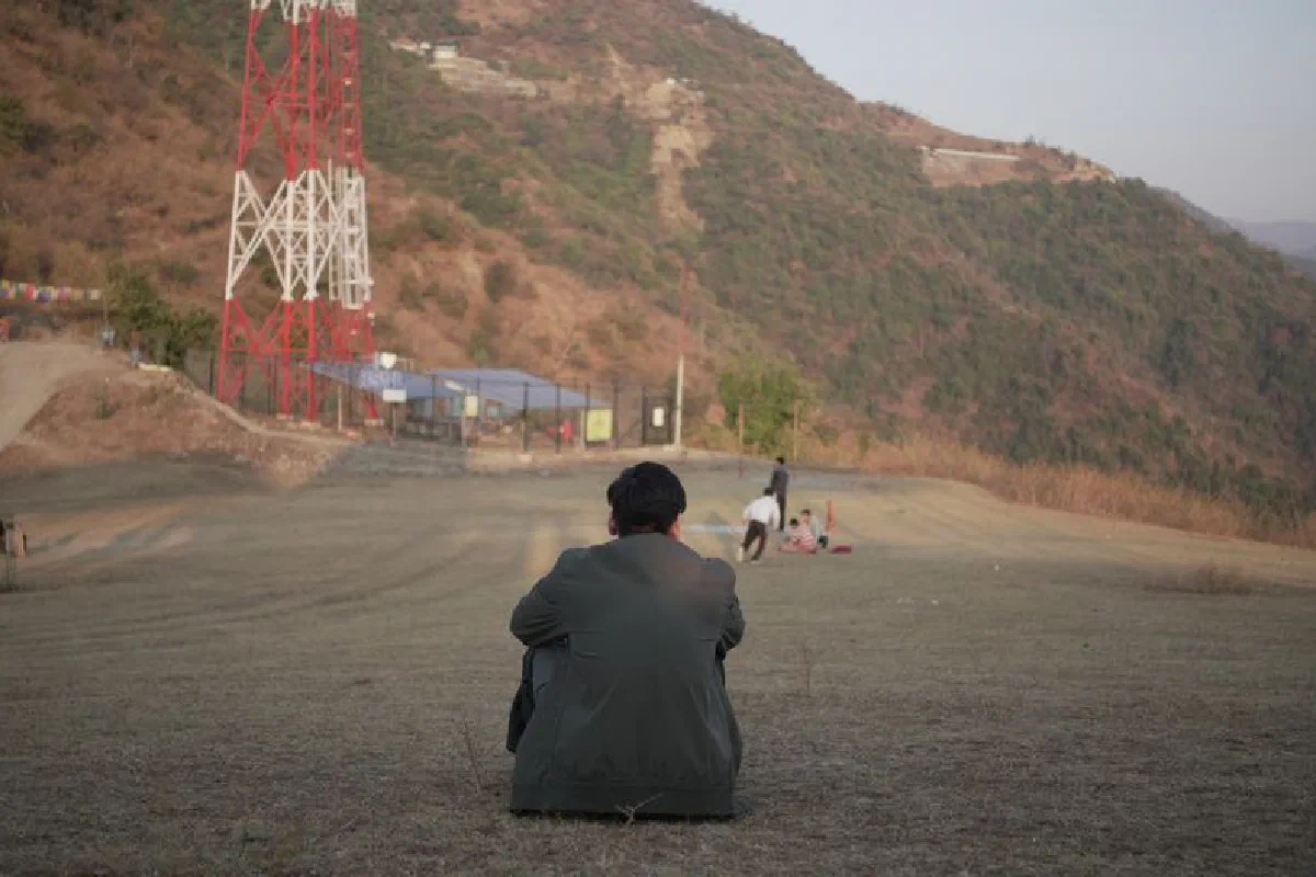 Man sits outdoors near a mountain with a red tower in view. Scenic landscape.