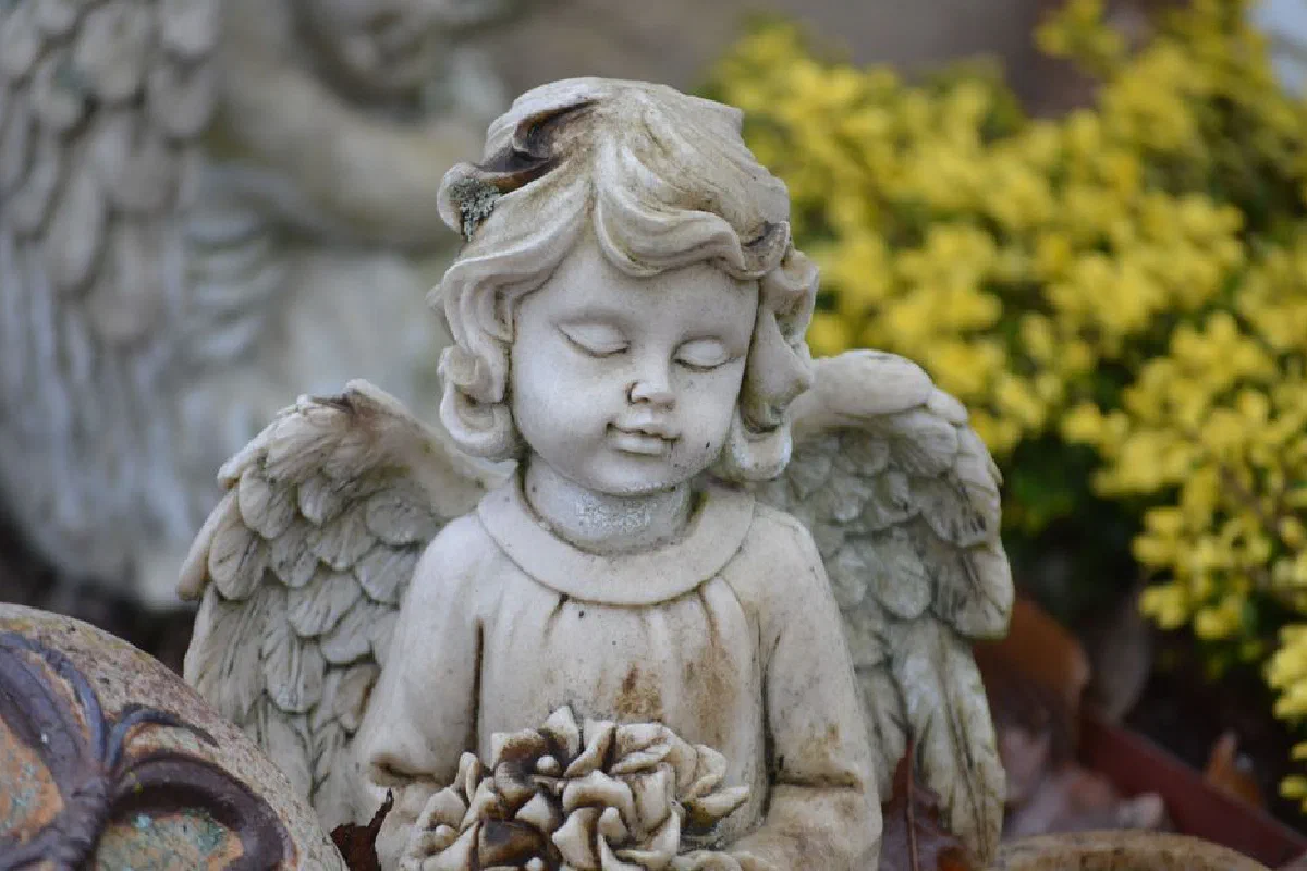 Close-up of a serene marble angel sculpture with wings in a garden with yellow flowers.