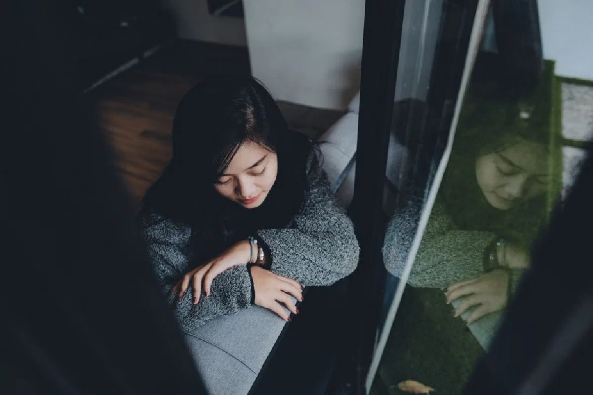 A young Indonesian woman in deep thought as she leans on a window sill indoors.