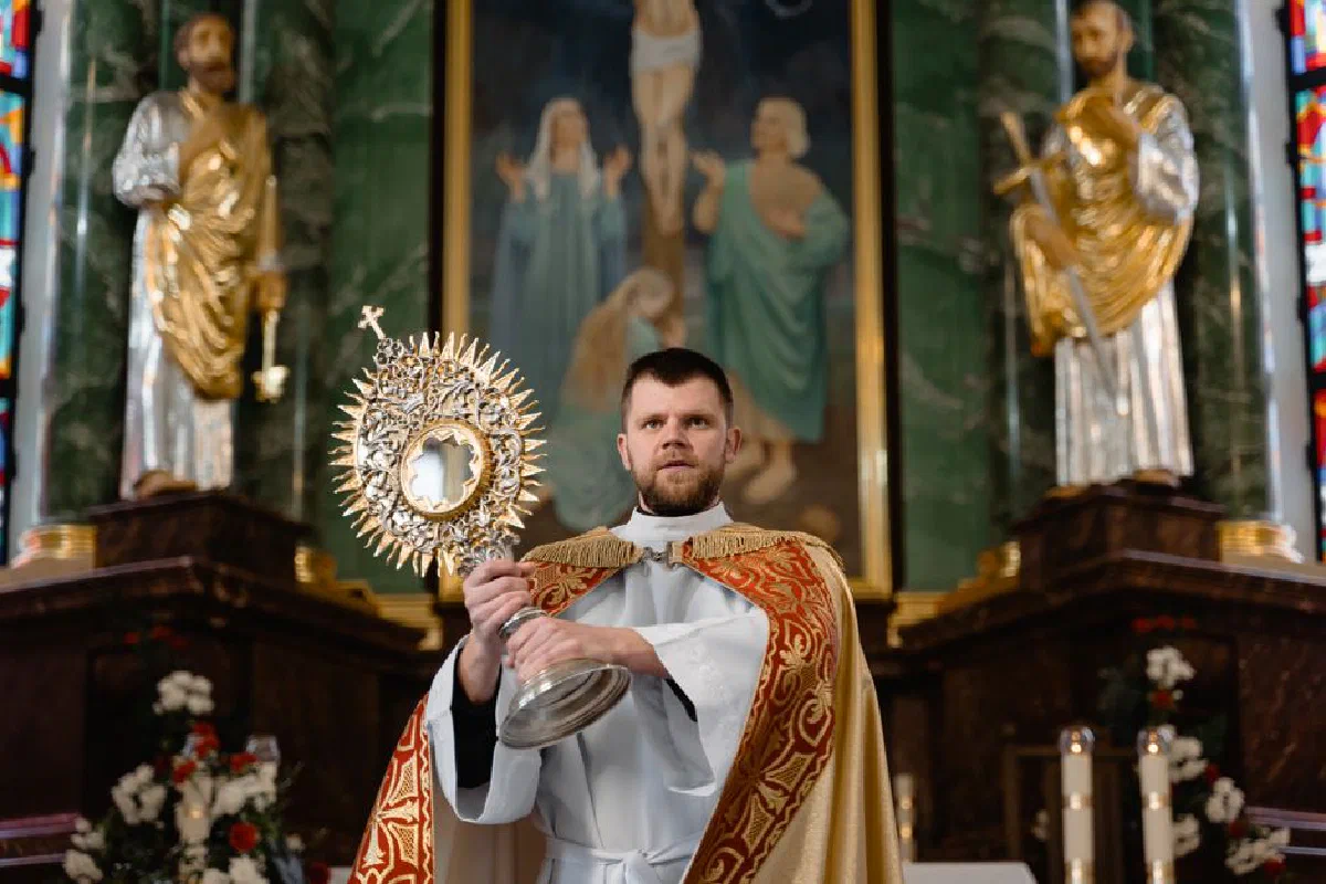 Priest in traditional vestments holding a monstrance during a religious ceremony inside a church.