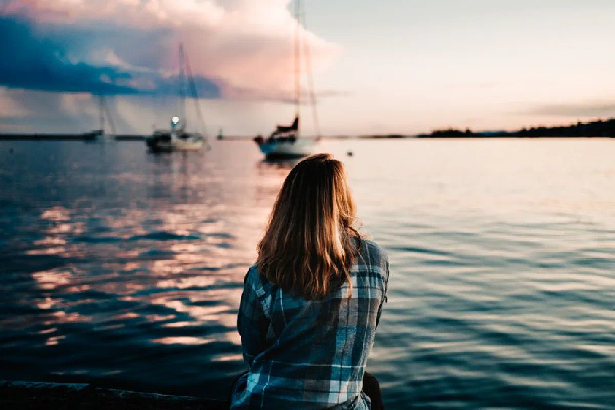Back view of lonely female sitting on seafront and looking away against blurred boats in bay at sunset
