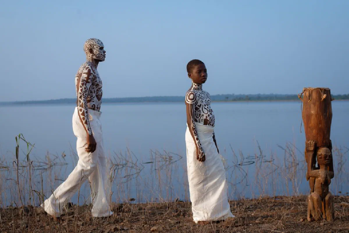 Two people with body paint stand by a serene lake, showcasing cultural art.