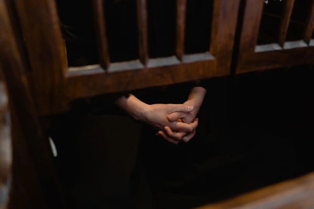 Hands clasped in prayer inside a wooden confessional, symbolizing faith and introspection.