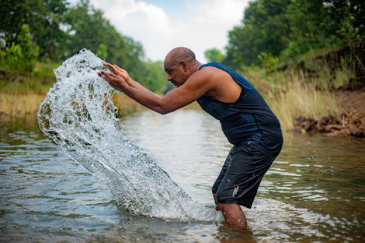 A man splashes water in a vibrant outdoor stream, surrounded by greenery.