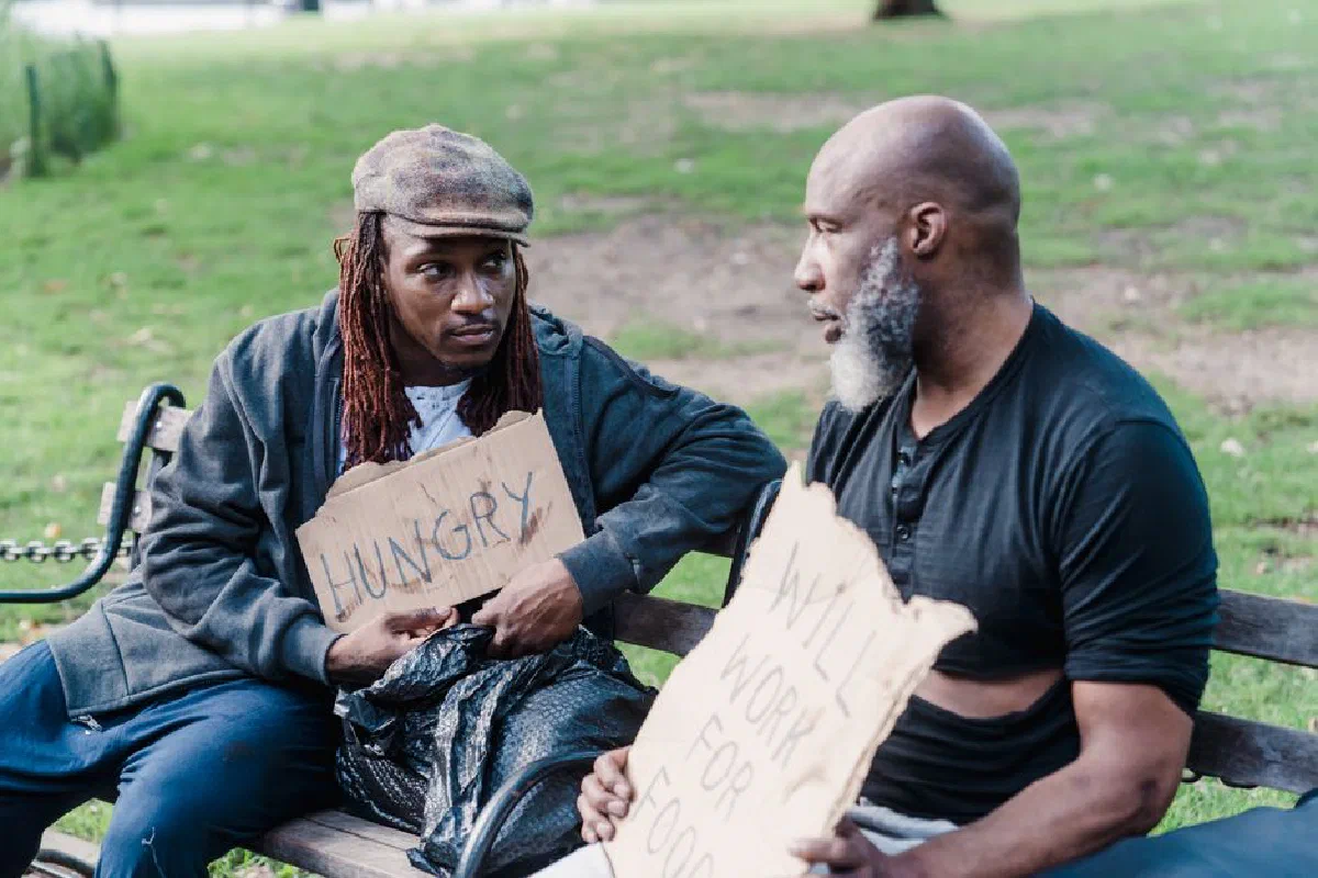Two men sitting on a bench in a park holding signs about hunger and work.