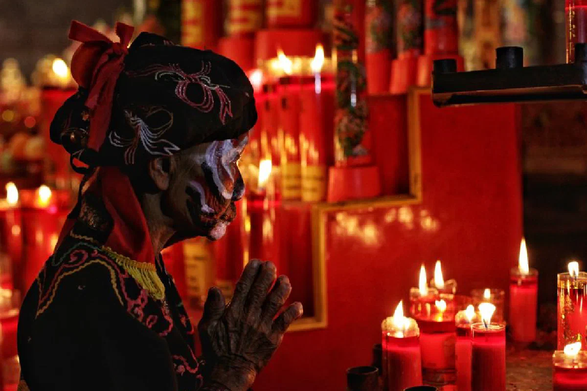 An elderly woman in traditional attire prays in a candlelit temple setting in Semarang, Indonesia.