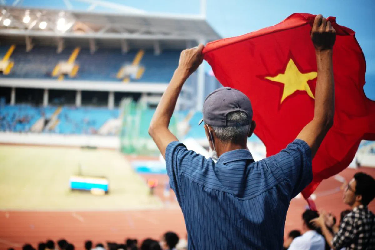 A man energetically waves a Vietnamese flag in a crowded sports stadium during the day.