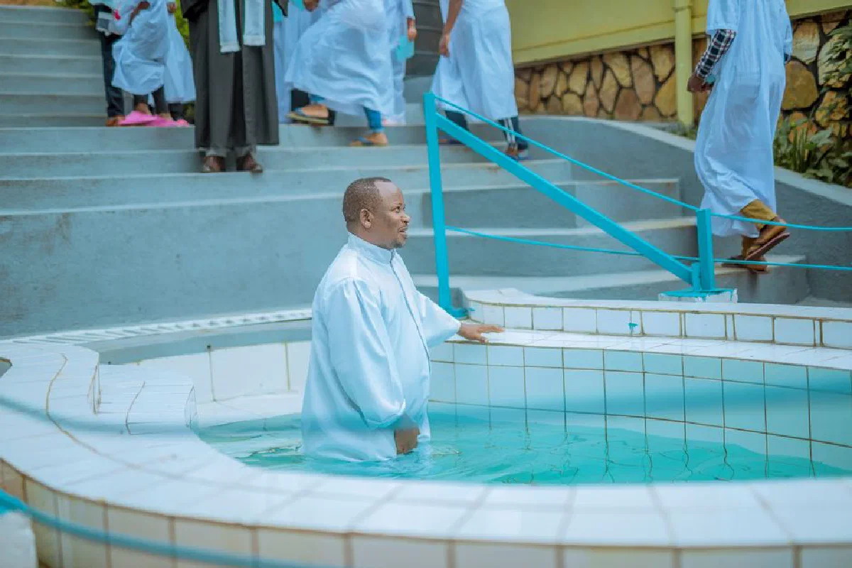 A serene outdoor baptism ceremony with a man in white attire in a tiled pool, surrounded by participants.