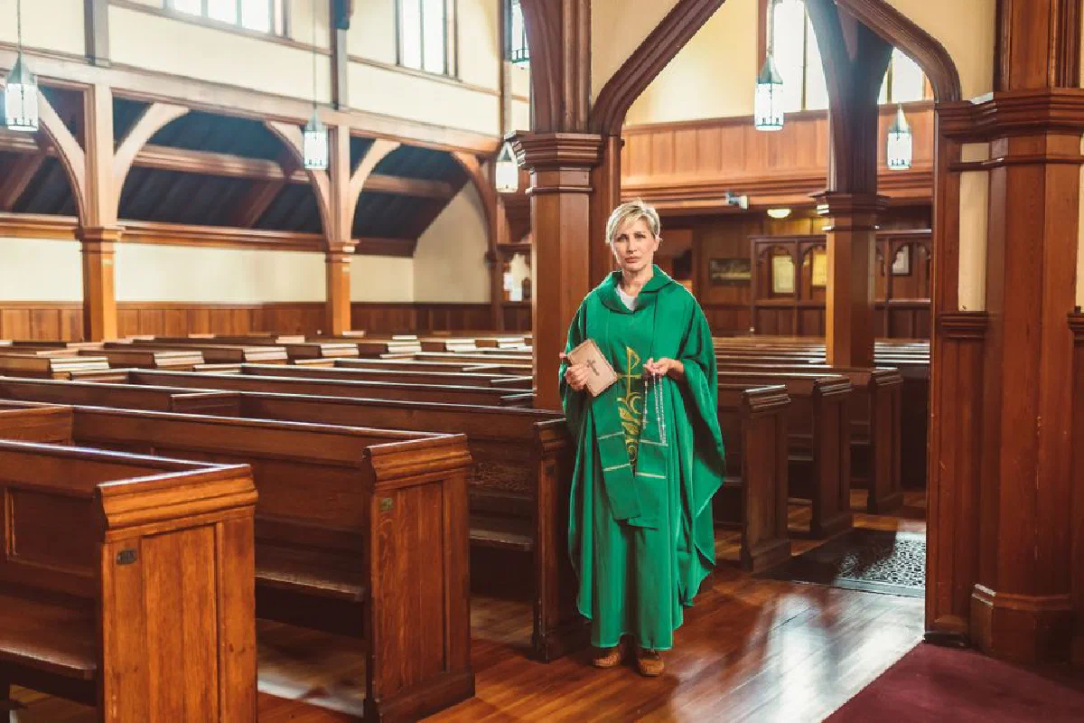 Woman priest in green robe standing among wooden pews of a traditional church interior.