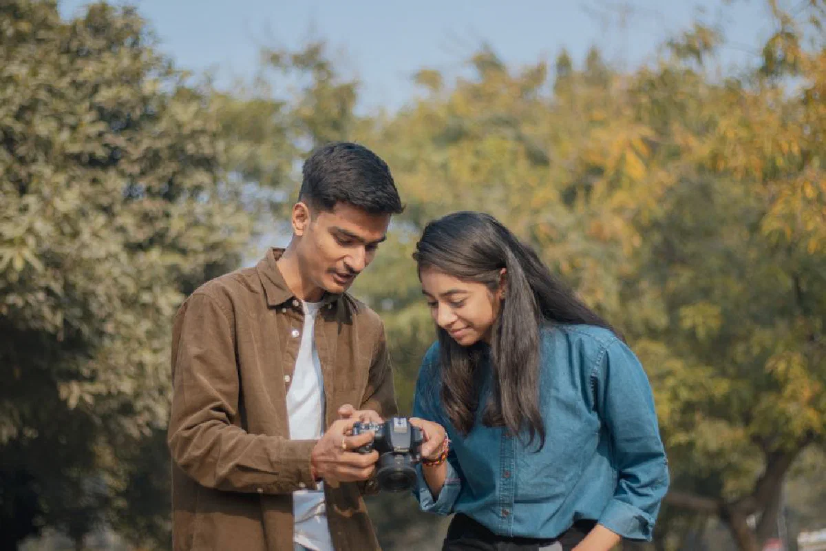 A young couple joyfully reviewing photos on a DSLR camera in a sunny park setting.