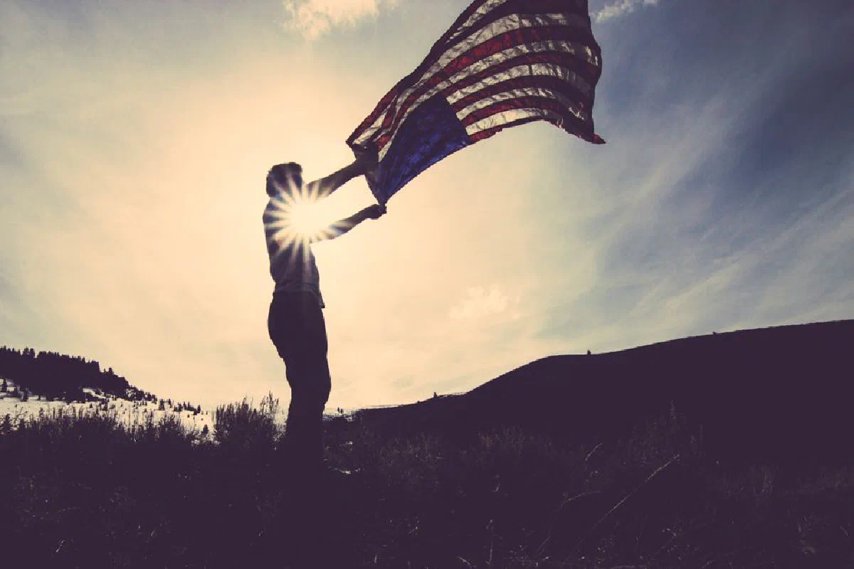 Silhouette of a person with an American flag against the bright sun in an open field.