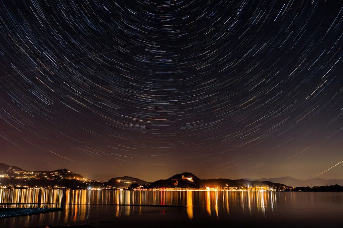 Star trails over a calm lake reflecting city lights, creating a serene and dramatic night scene.