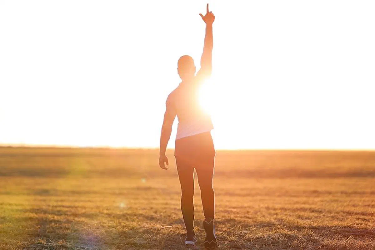 A silhouette of a man with raised hand pointing to the sky in an open field at sunset.