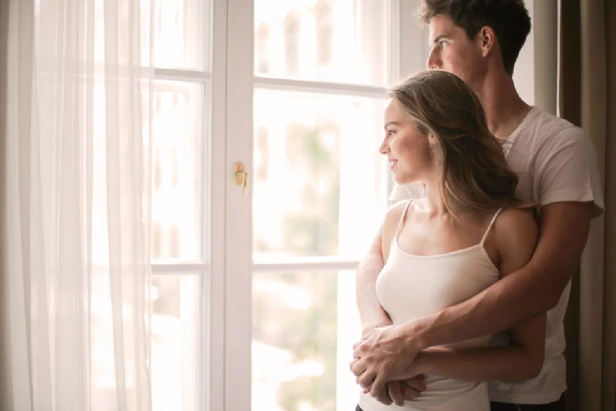 Side view of content young couple in casual clothes standing and hugging while enjoying weekend and looking out of window together