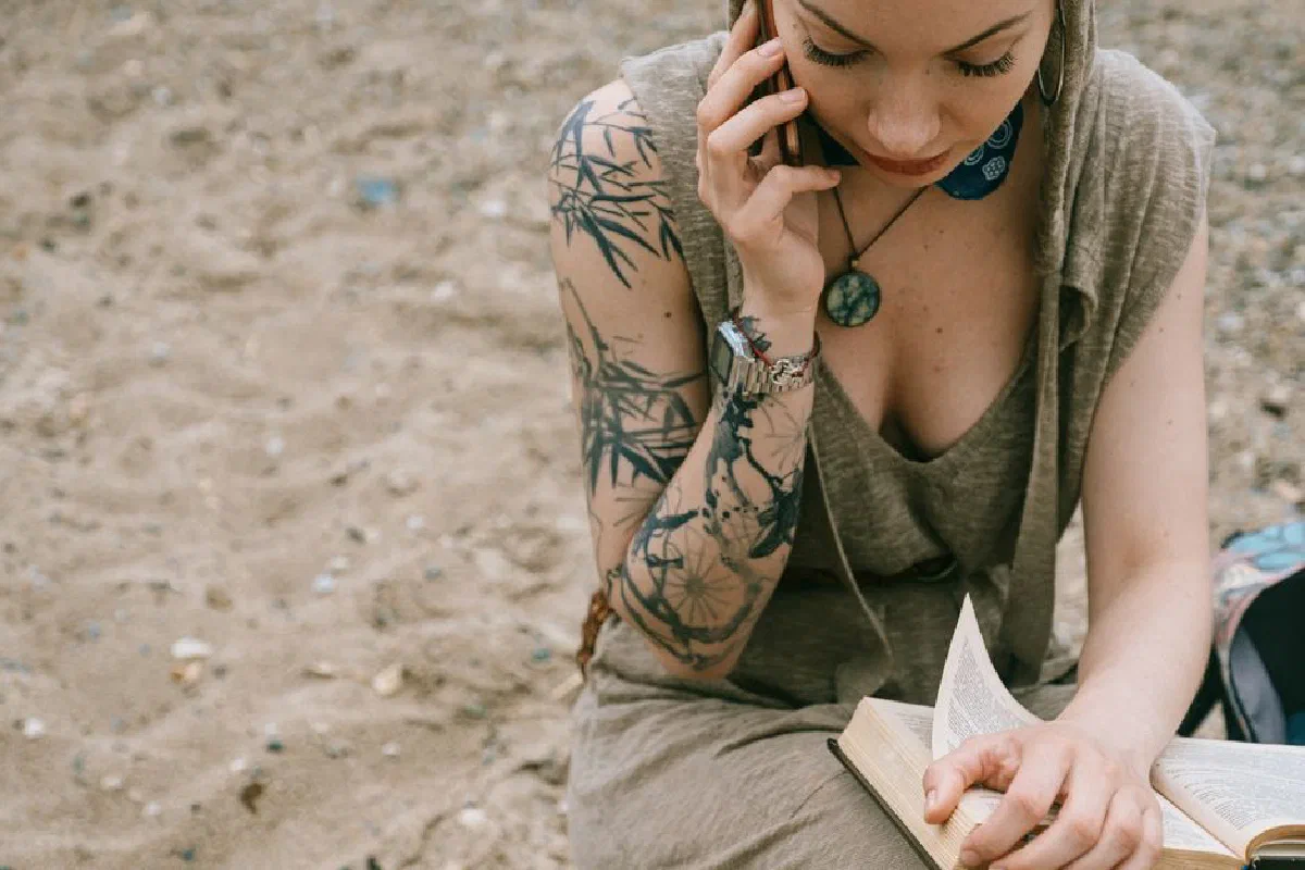 A woman reads a book while talking on her phone at the beach.