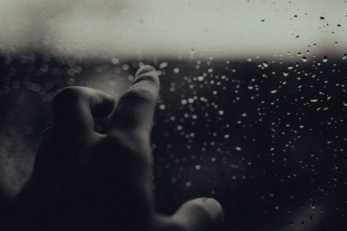 A hand reaches out to a rain-streaked window, creating a contemplative mood in monochrome.