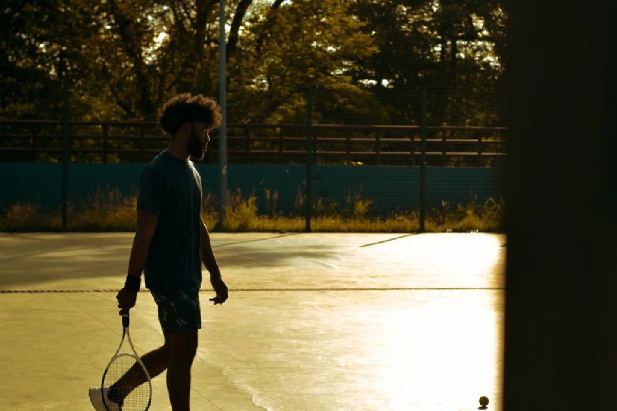 Man walks on a sunlit tennis court at sunset, holding a racket. Perfect lighting for sports imagery.