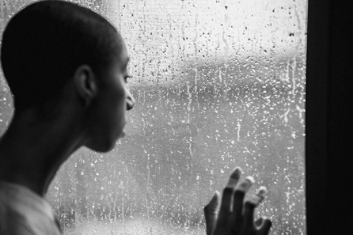 Black and white of melancholy androgynous African American female with short hair standing near wet window at rainy weather and looking away in daytime