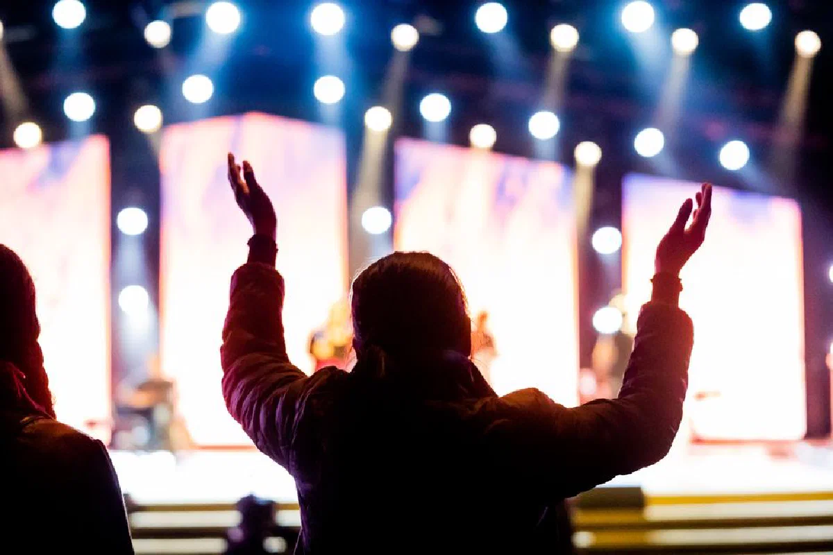 Silhouette of person worshiping with raised arms at a bright music concert stage.