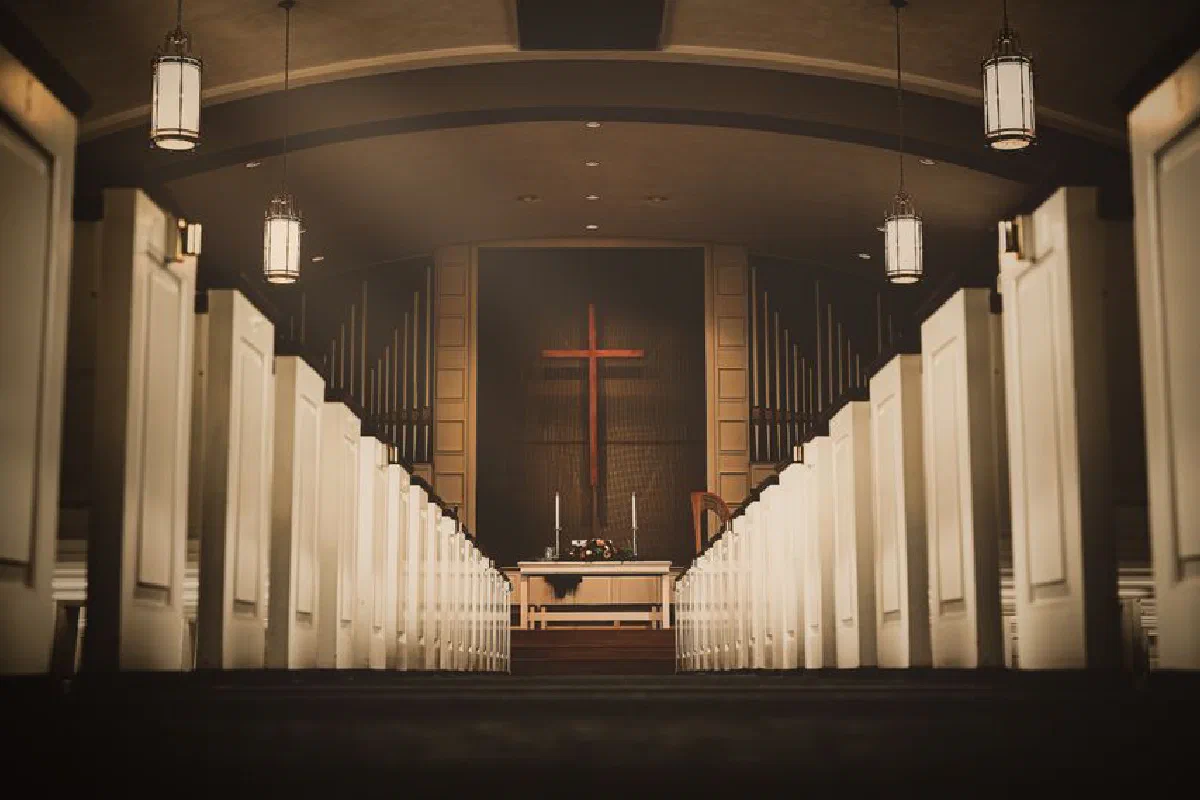 A beautiful church interior with a central aisle leading to a cross and altar, illuminated by hanging lights.