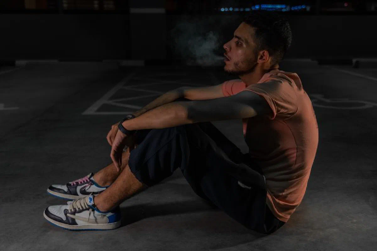 A young man in casual clothing sitting on the ground in a dimly lit urban setting.