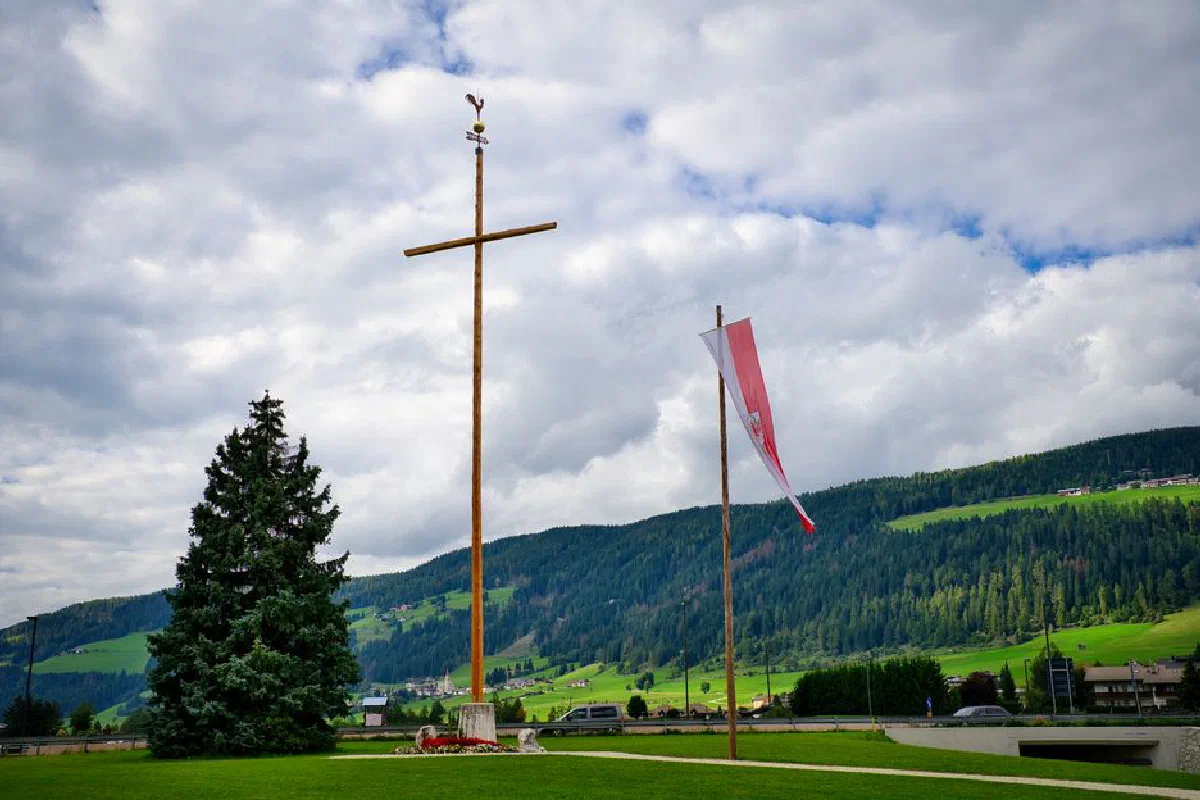 Cross and flag in Toblach, Italy countryside with lush green hills and cloudy sky.