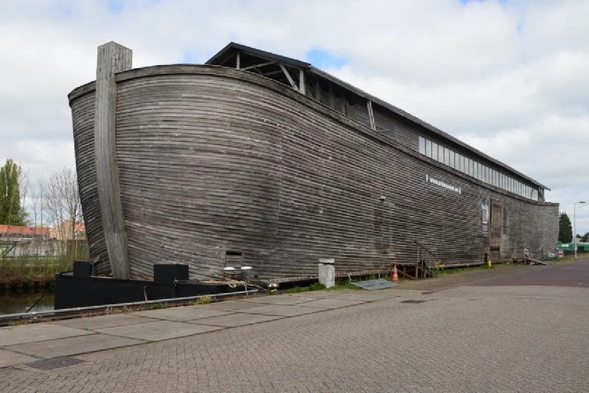Large wooden structure by canal, resembling a historical ark on a cloudy day.