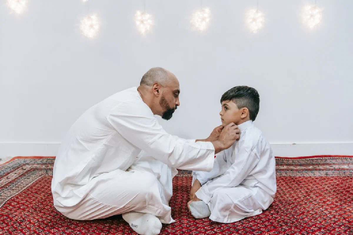A father gently adjusting his son's collar, sitting on a prayer mat indoors.