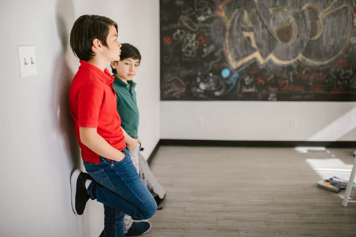 Two boys leaning against a wall in a classroom with graffiti board.