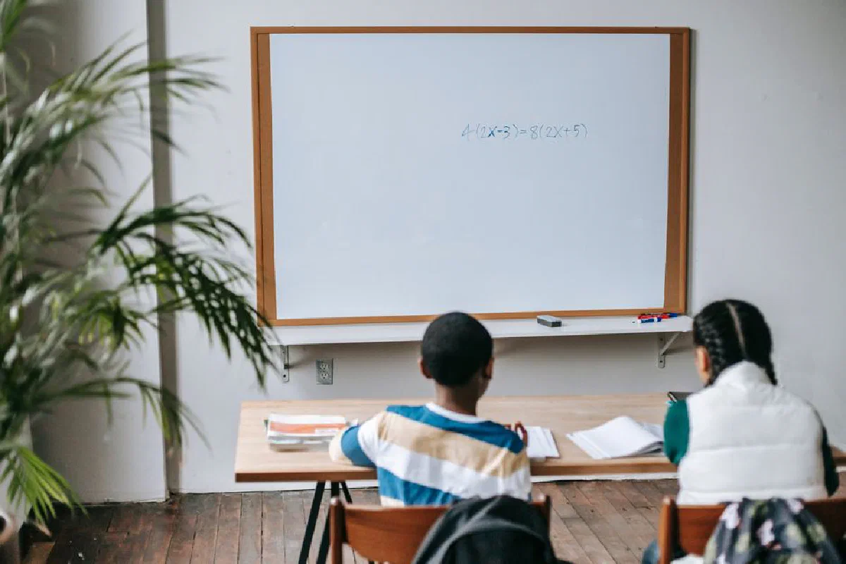 Back view unrecognizable multiethnic pupils sitting at desk with copybooks against whiteboard in light modern classroom