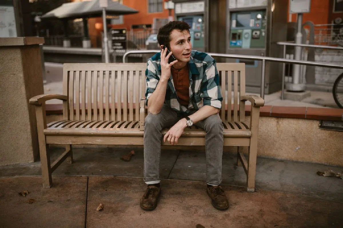 A young man in plaid shirt talks on phone while sitting on a wooden bench outdoors.