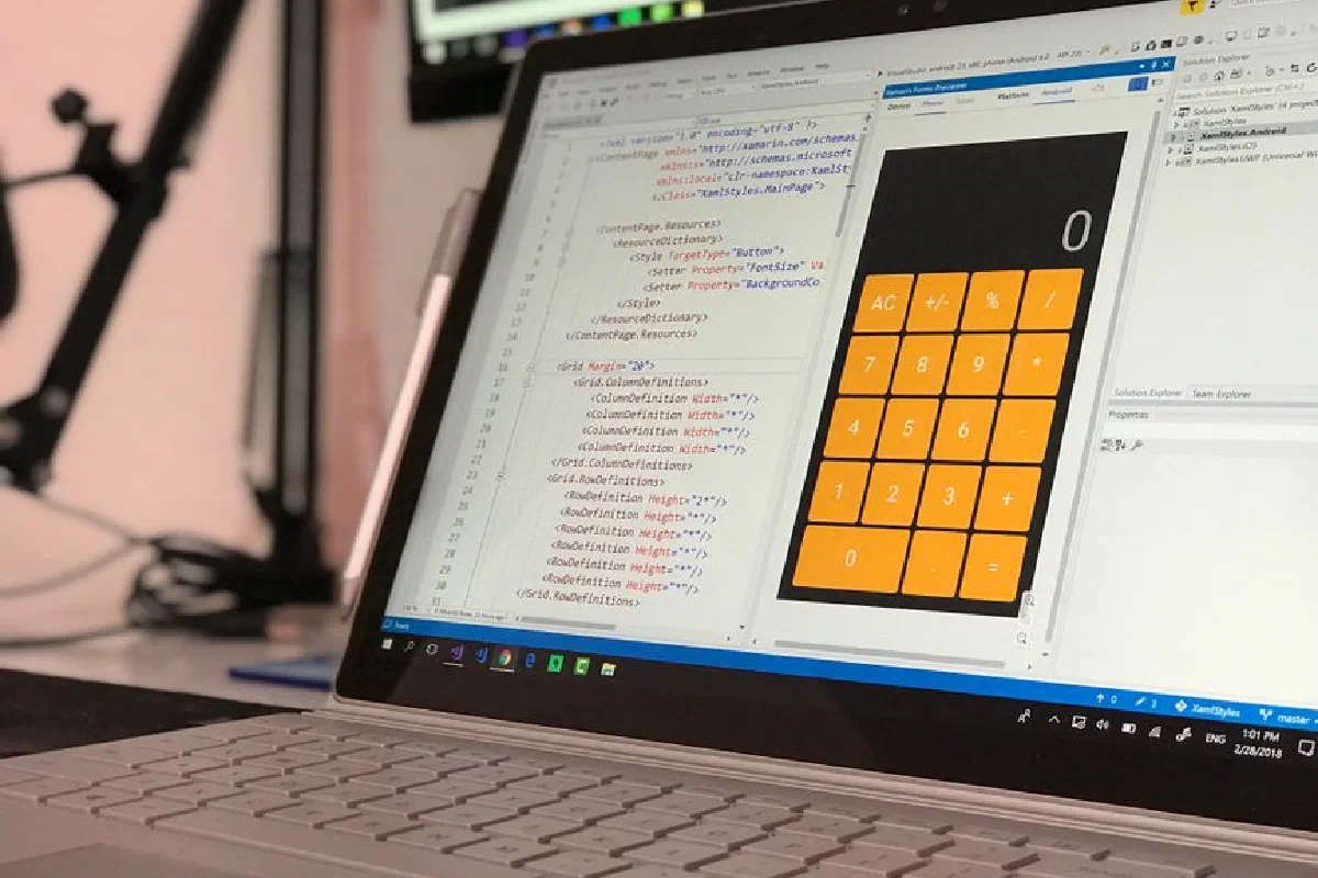 A laptop screen shows a coding application with a calculator design in a tech office setting.