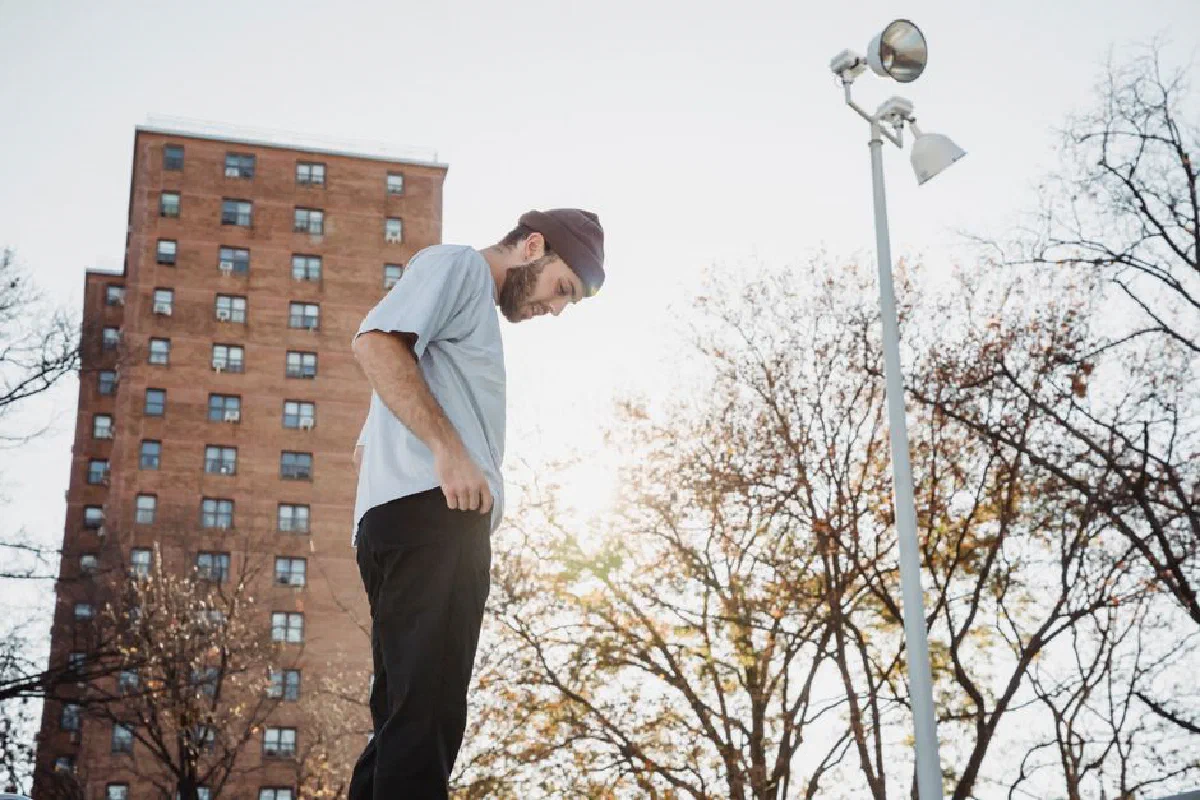 From below side view of young unshaven male in informal apparel looking down against urban building and trees in fall