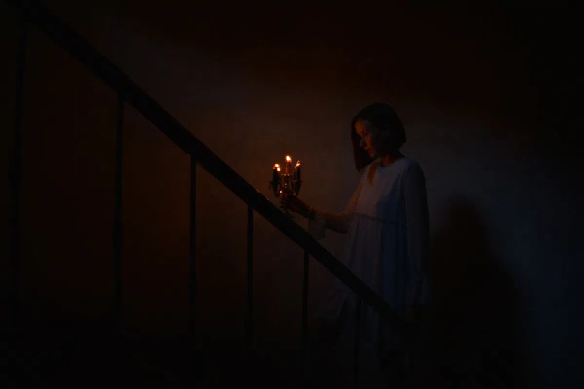 A woman in a nightgown ascends dimly lit stairs holding candles, creating a mysterious ambiance.