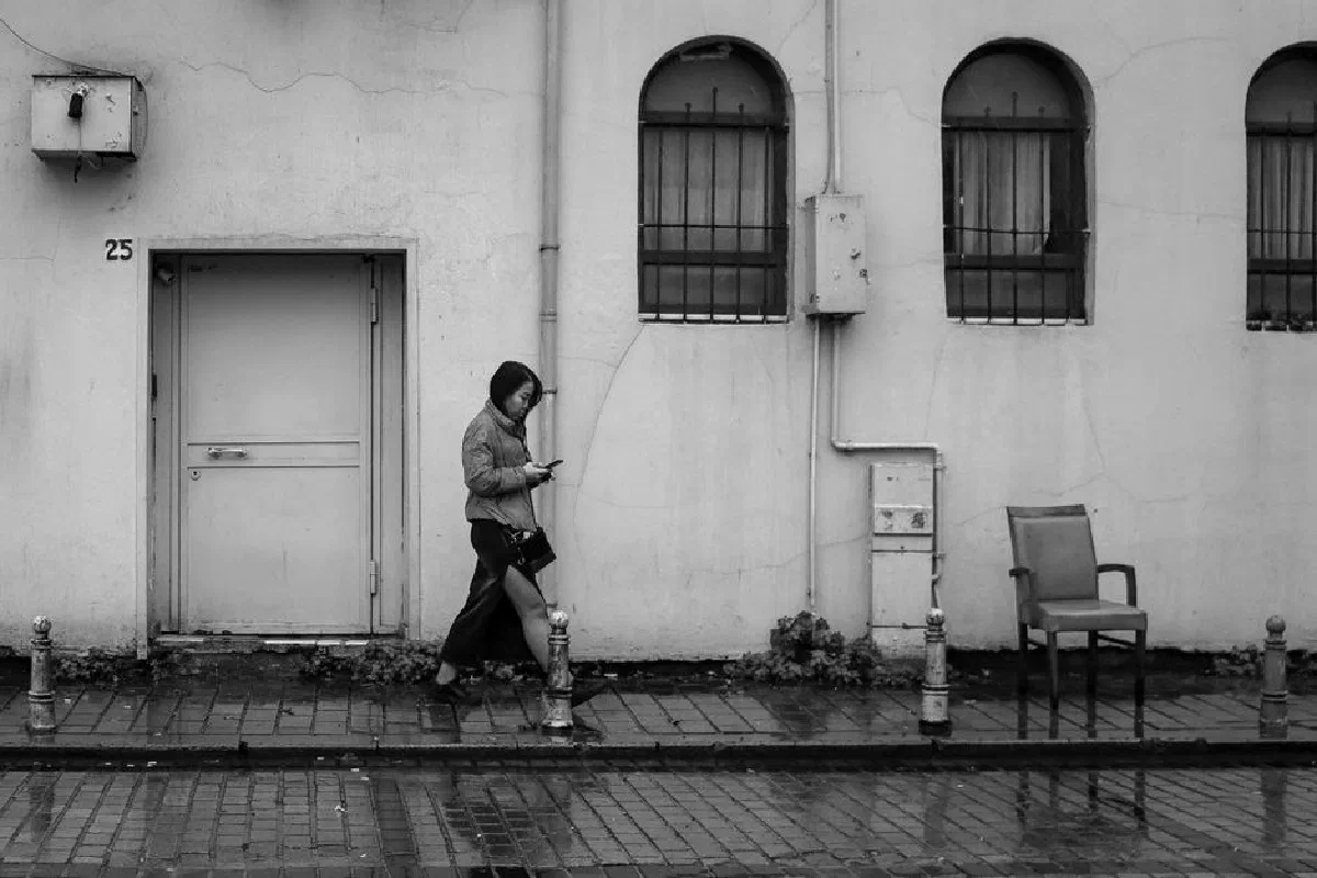 A woman in a hoodie walks on a wet sidewalk using a smartphone. Black and white urban scene.