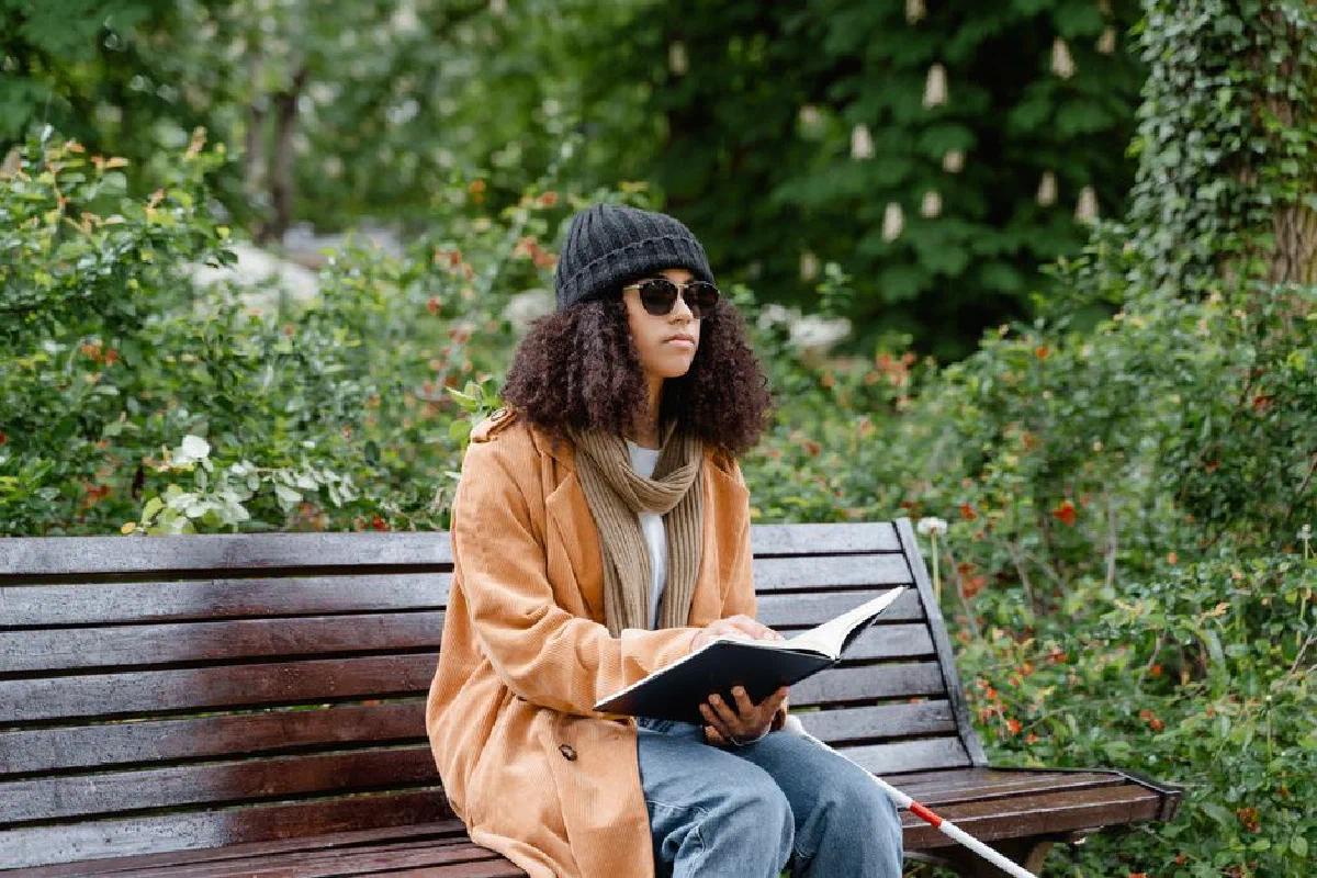 A young woman wearing sunglasses and a beanie reads a braille book while seated on a park bench.