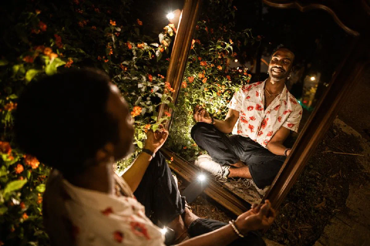 A man meditates with a reflection in a mirror outdoors, surrounded by vibrant flowers during nighttime.