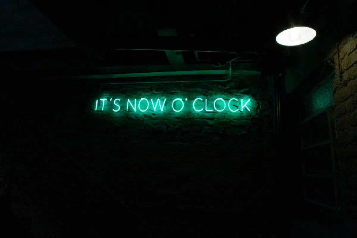 Green neon sign in a dimly lit room showcasing 'It's Now O' Clock'.