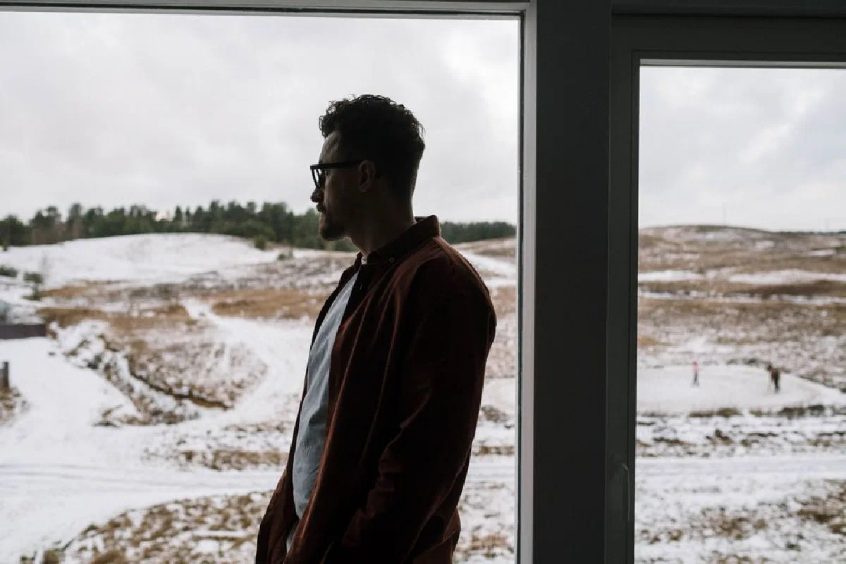 A man in deep thought gazes through a window at a tranquil snowy landscape.