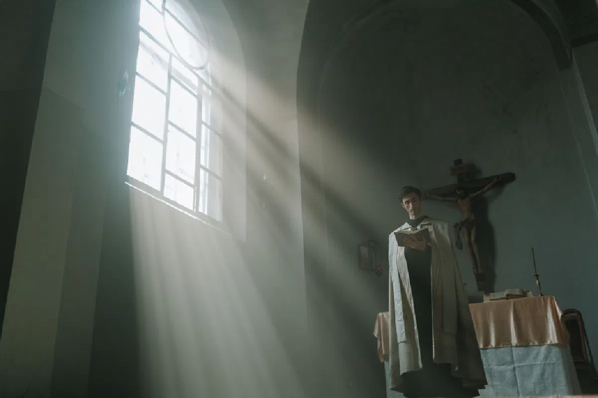 A priest in a church reads under dramatic sunlight with a crucifix in the background.